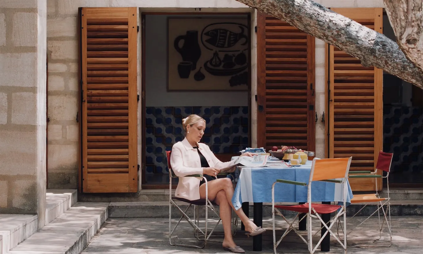 Kvinne i en hage leser ved et bord med blå duk, omgitt av stoler og åpne trepersienner.