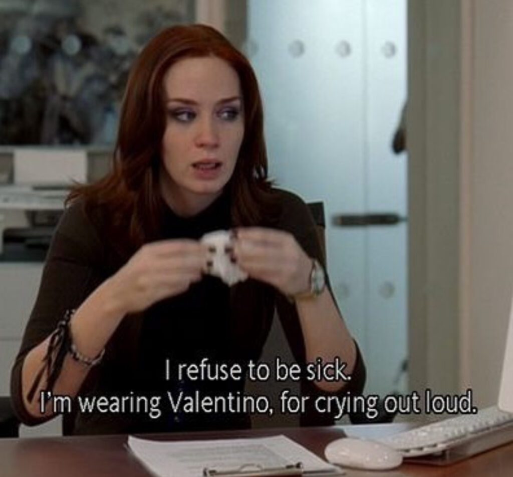 Kvinne ved skrivebord ser bekymret ut, holder et lommetørkle, med tekst: "I refuse to be sick. I'm wearing Valentino, for crying out loud."