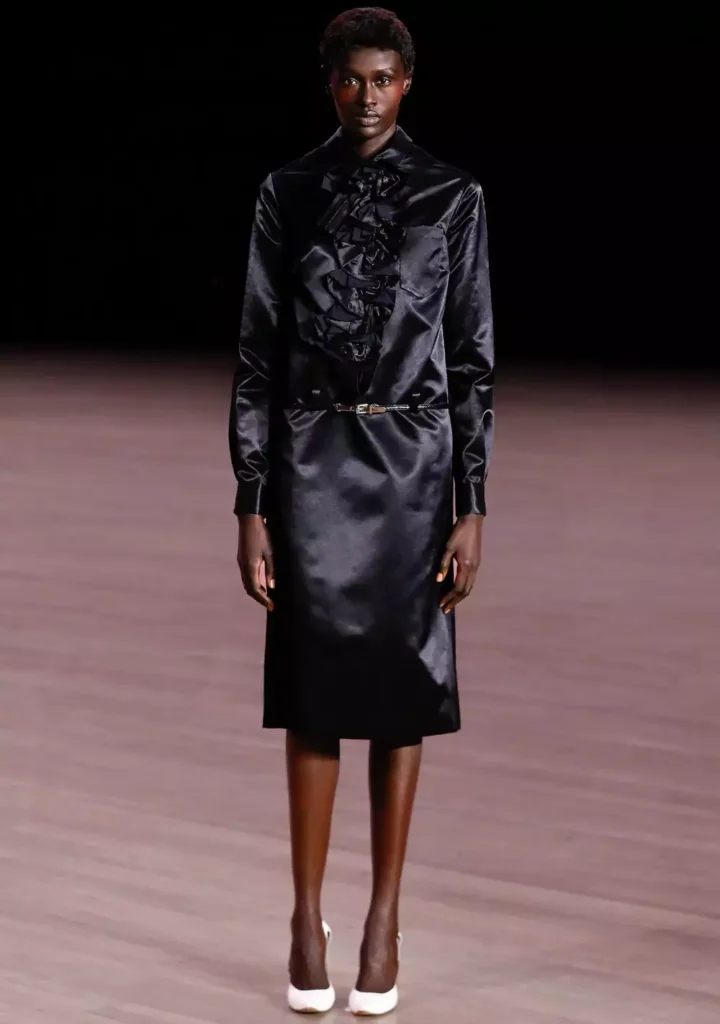 Modell i elegant svart kjole med rysjer på catwalken, iført hvite sko.